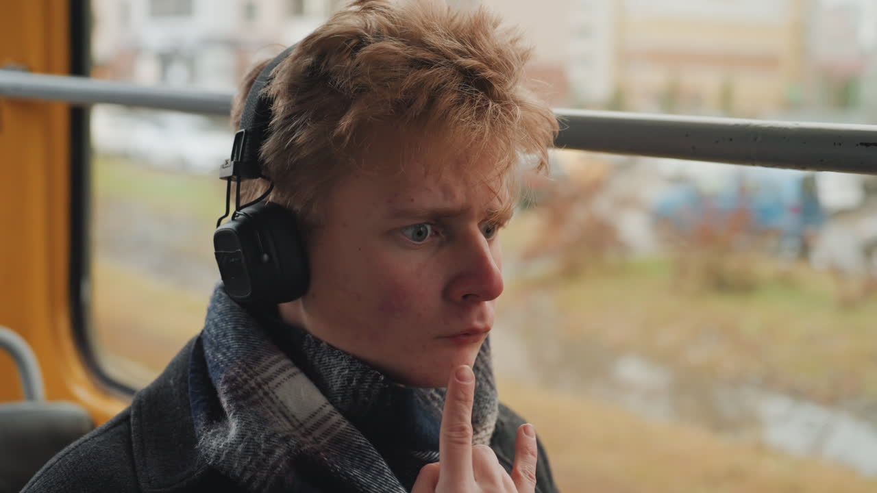 Weißer männlicher Künstler in der Straßenbahn, Kopfhörer hörend und aus dem Fenster blickend, in Schal und Mantel gehüllt. Nachdenkliche Handgesten deuten auf das Entwickeln von Ideen hin, während eine bewölkte Stadtlandschaft vorbeizieht, filmisch.