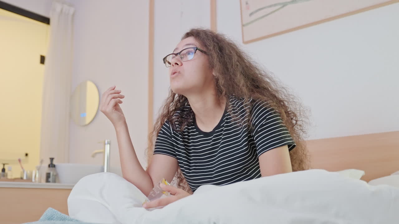 Giovane donna seduta sul letto dell'hotel che sorride mentre prende uno snack e guarda verso la finestra. Occhiali e capelli ricci incorniciati dalla lampada da comodino e dalla toeletta, routine mattutina rilassata durante il soggiorno in hotel.