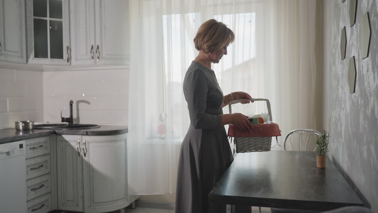 Back view of house owner walking into bright kitchen with gift basket picking item from basket placing it on dark table modern cozy interior with stainless plate on counter