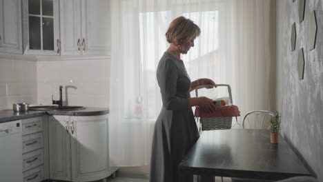 back view of house owner walking into bright kitchen with gift basket picking item from basket placing it on dark table modern cozy interior with stainless plate on counter