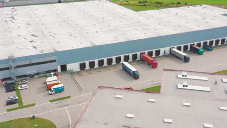 Aerial-following-shot-of-semi-trailer-trucks-travelling-along-a-warehouse-on-the-parking-lot-in-the-logistics-park-with-loading-hub