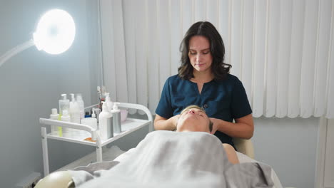 cosmetologist massaging head of client then retrieving cotton pad to dip into purified water during facial therapy session in bright spa room with medical lamp and skincare products nearby