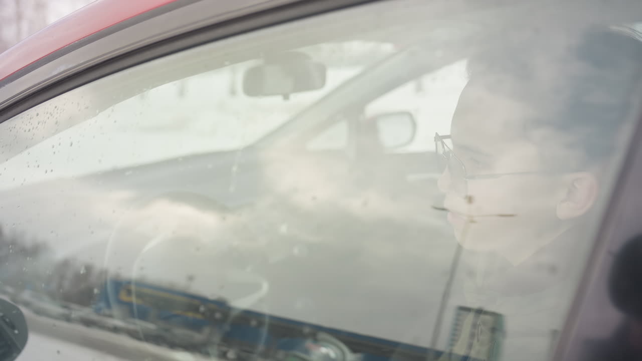 young adult wearing sunglasses sits in driver seat during winter day, slowly rolls up car window while holding steering wheel and gazing outside with thoughtful expression