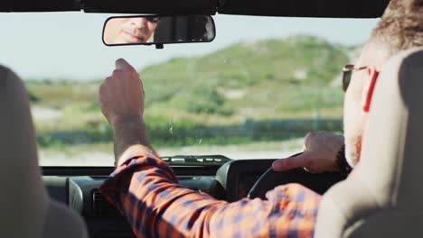 hombre caucásico con gafas de sol sentado en el coche ajustando el espejo retrovisor en un día soleado en la playa