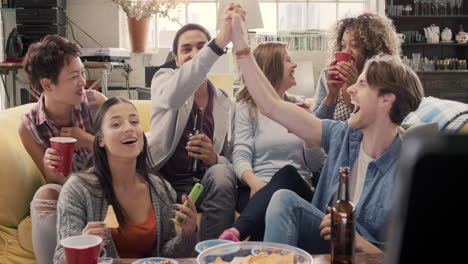 diverse group of student friends arms raised celebrating goal watching sports event on tv together eating snacks  drinking beer