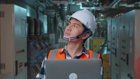 close up of asian male engineer with safety helmet working on a laptop and looking around while standing in engine control room, work of electrical generators