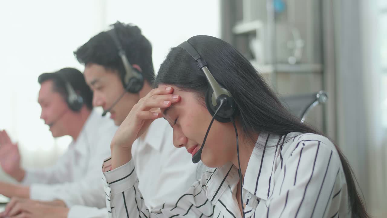 A Woman Of Three Asian Call Center Agents Wearing Headset Is Tired Due To Working While Two Of Her Colleagues Are Speaking And Typing During The Call With Customer At The Office