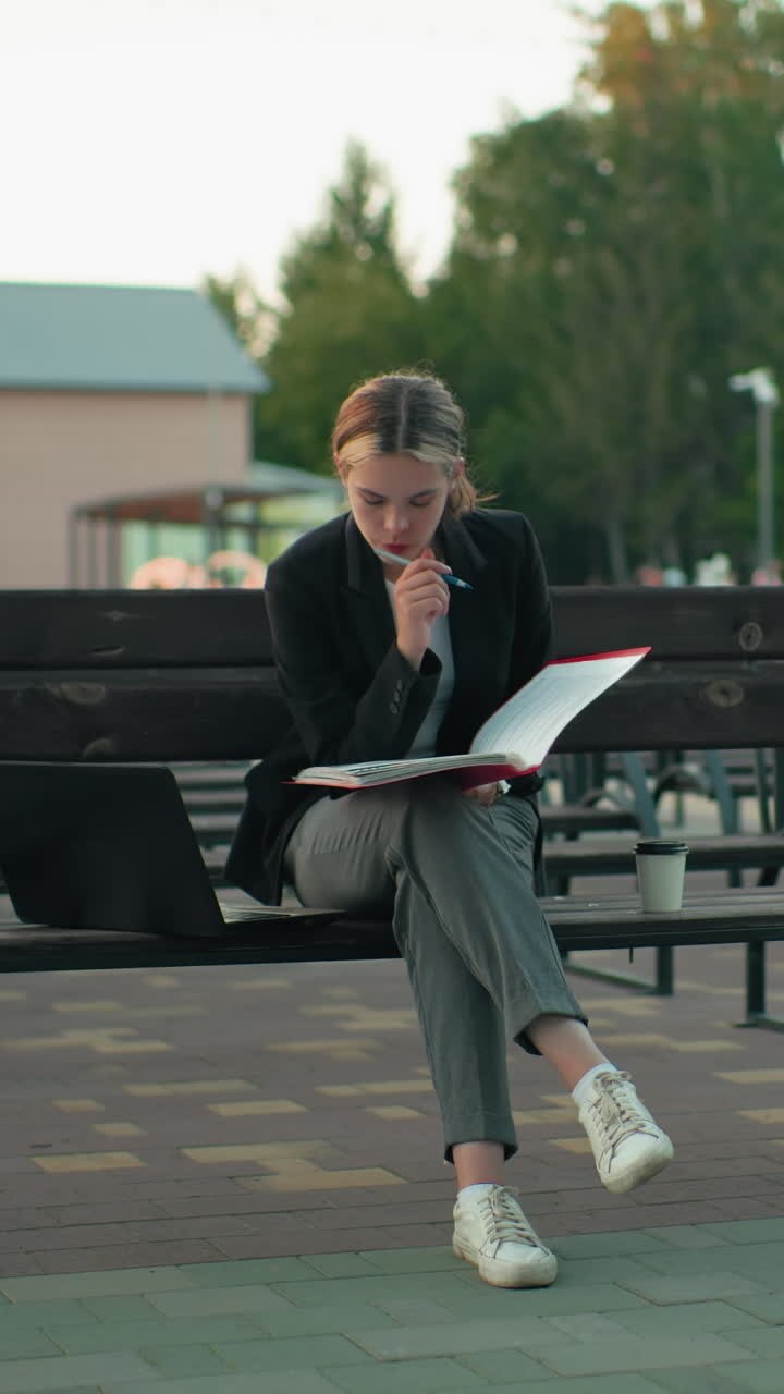 Ung dame sitter alene på en utendørs benk, dypt konsentrert mens hun leser en stor, åpen bok. Hun holder en penn, og har en laptop og en kaffekopp ved sin side, i rolige, urbane omgivelser omgitt av tomme benker