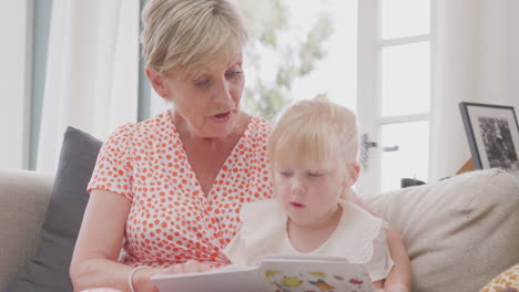 abuela sentada en el sofá con su nieta en casa leyendo un libro juntos