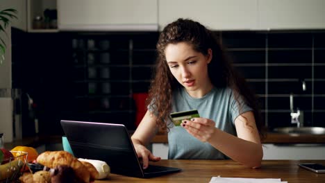 joven alegre haciendo compras en línea usando una computadora portátil y una tarjeta de crédito mientras desayuna en la cocina de casa