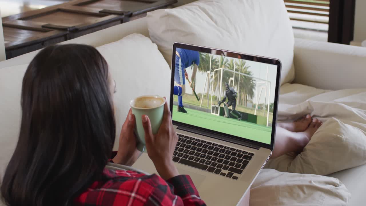 Composite of woman sitting at home holding coffee watching hockey match on laptop