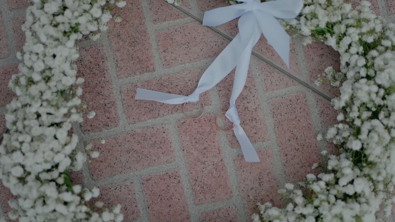 Two wedding rings tied with white ribbon on a baby's breath wreath over brick pavement