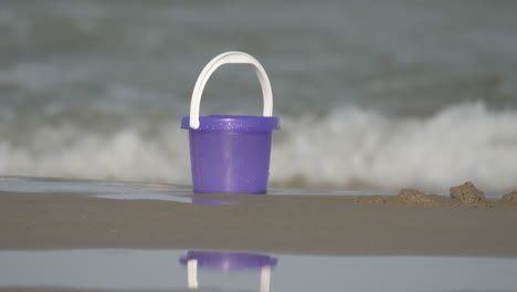 a purple toy bucket on the sand on the beach with sparkling waves on the background