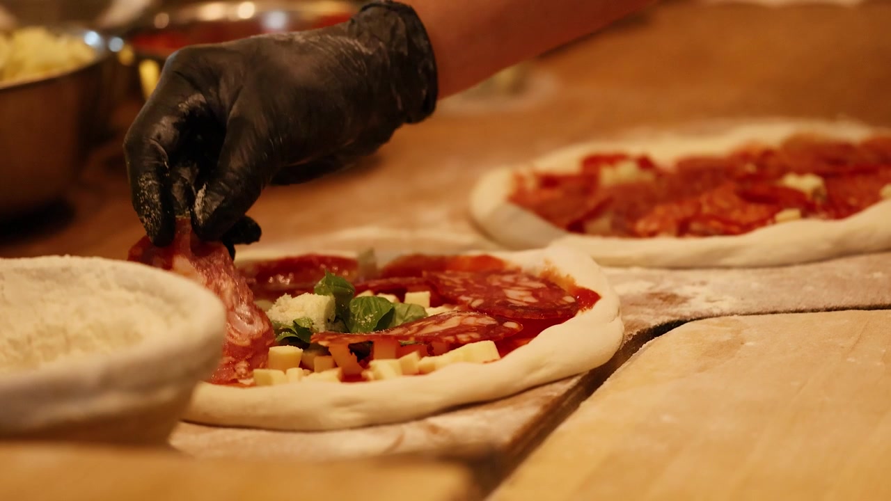 Primo piano delle mani dello chef che posizionano il pepperoni sull'impasto crudo della pizza in una cucina professionale.