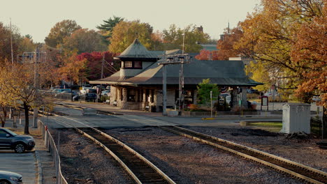 Coches-Que-Cruzan-La-Intersección-Sobre-Las-Vías-Del-Tren-Con-La-Estación-De-Tren-De-Fondo-En-Una-Hermosa-Tarde-De-Otoño-A-La-Hora-Dorada-En-Kirkwood-En-St.