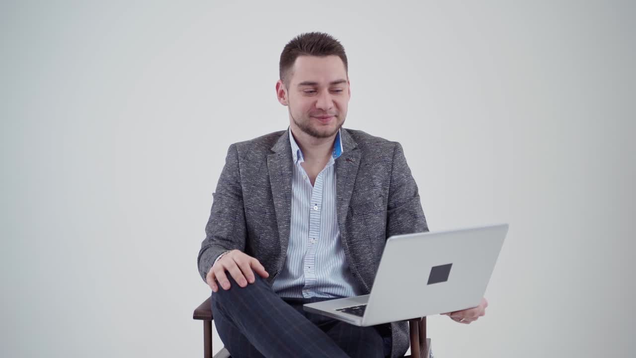Portrait of a handsome businessman with a laptop. Smiling young man in grey costume sitting on the chair on the white background. Slow motion.