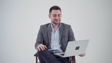 portrait of a handsome businessman with a laptop. smiling young man in grey costume sitting on the chair on the white background. slow motion.
