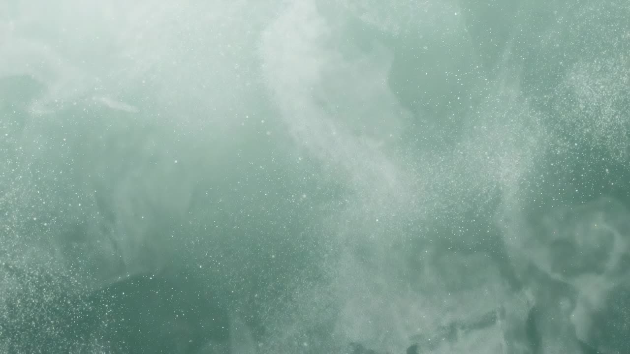 Forming teal-green mist plume brightening in contained fluid medium, fine white specks clustering