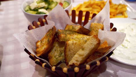 Mit-Knoblauch-Gewürzte-Croutons-Liegen-In-Einem-Stilvollen-Korb,-Mit-Griechischem-Salat-Im-Hintergrund