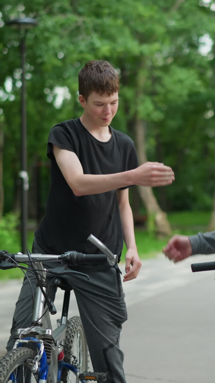 두 명의 친구가 자전거를 타고, 회색 을 입은 사람은 친구가 그를 환영하기 위해 올라오는 것을 기다리고, 회색을 입은 소년은 손목 부히기 위해 손을 고, 배경에는 흐릿한 푸른 나무가 있습니다.