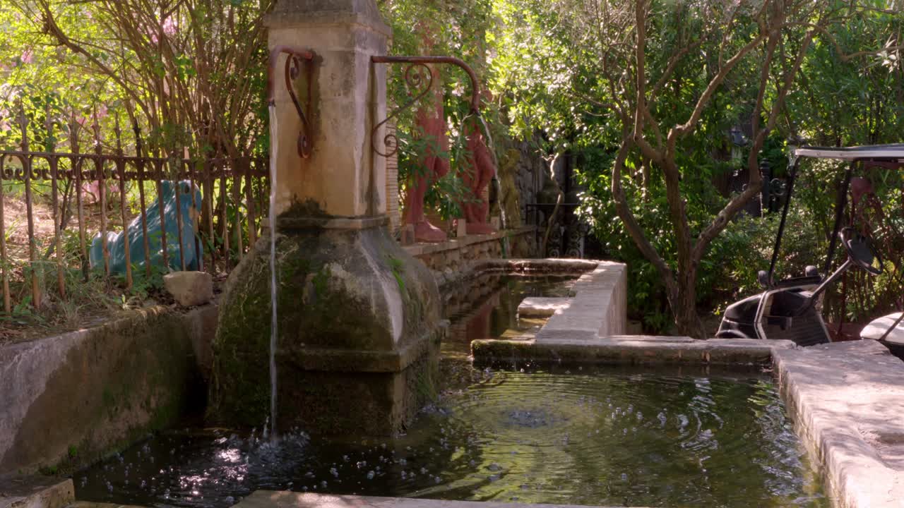 wide shot of a rustic stone fountain in Provence, France, with a rusted metal pipe faucet dispensing water into a small pool, surrounded by greenery, a golf cart, and a stone wall with statues.