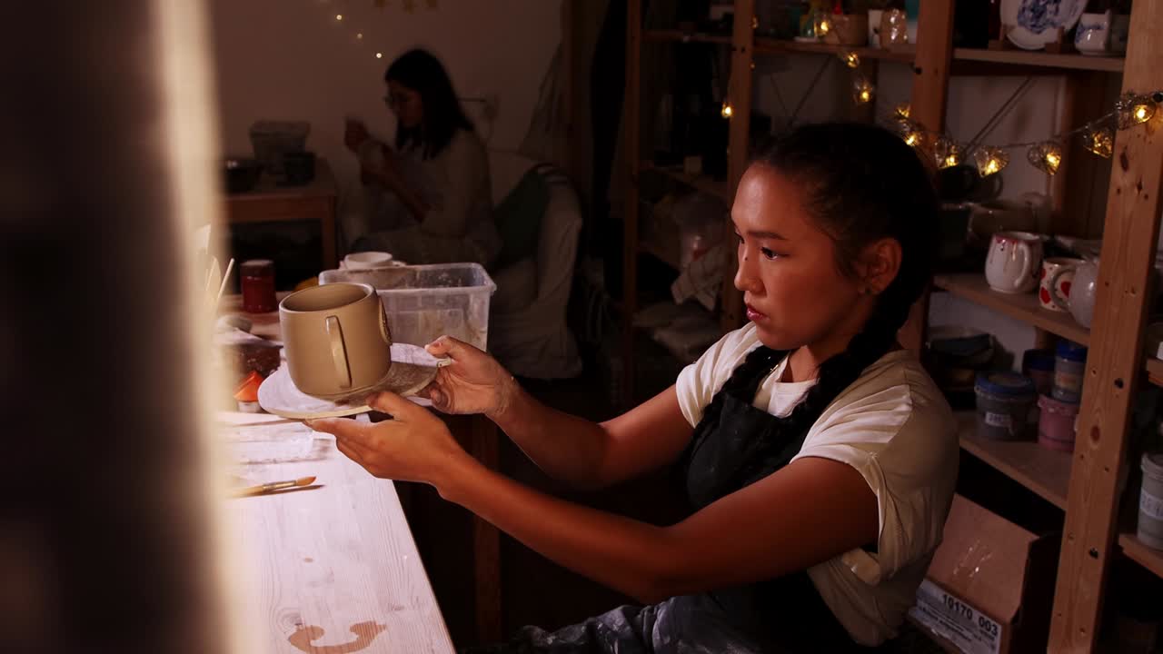Ceramica in studio - una vasaia applica il manico a una tazza e la osserva sotto una luce calda.