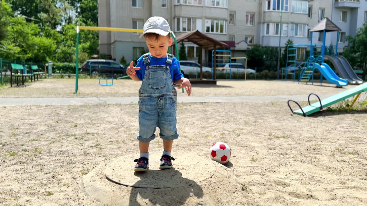 One year old toddler wearing jeans and cap is at the playground. Baby boy touching sand covering the ground.