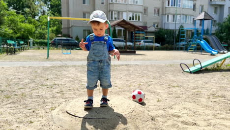 one year old toddler wearing jeans and cap is at the playground. baby boy touching sand covering the ground.