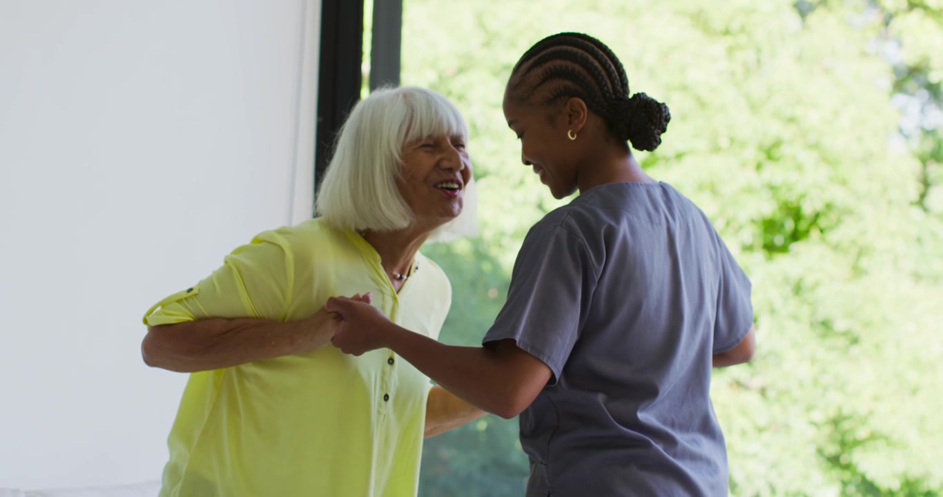 African American caregiver in scrubs taking hands lifting senior from sofa walking supporting walk