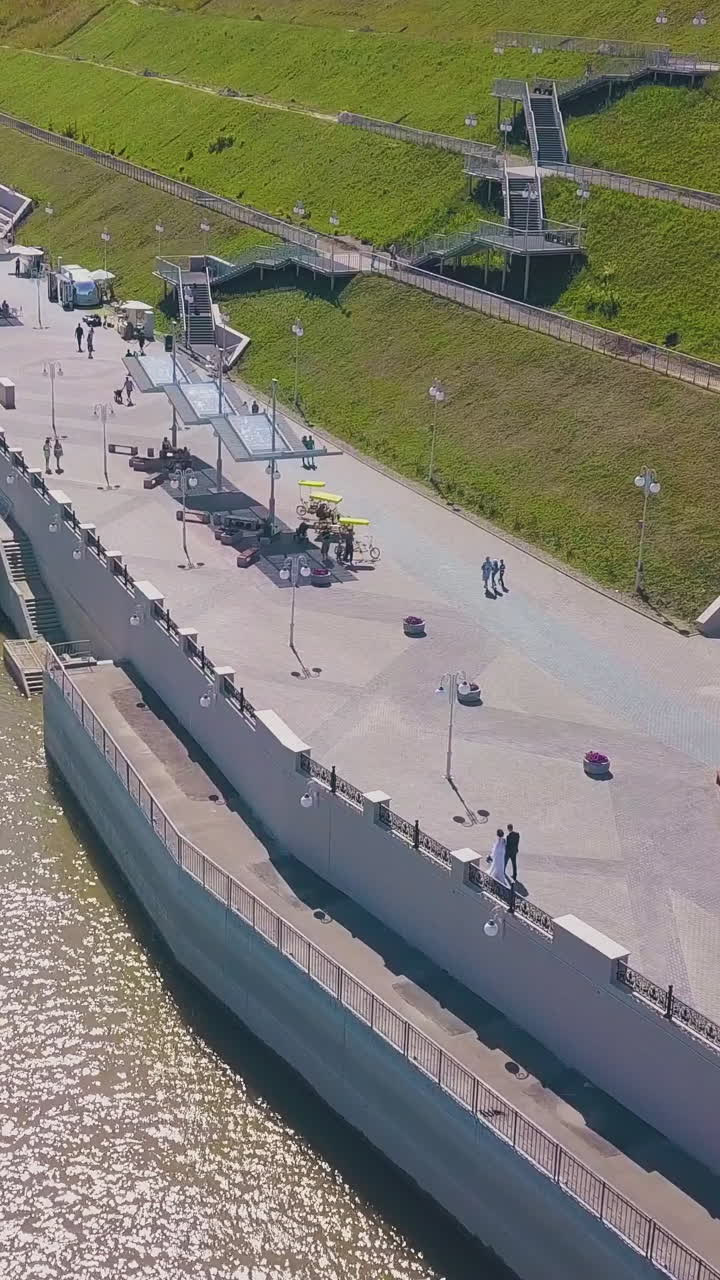 distant newly wedded couple walks along wide embankment at calm river and green hilly bank on sunny day bird eye view