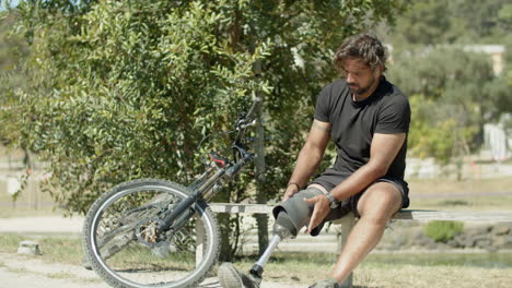 un tiro largo de un atleta serio arreglando una pierna protésica en el parque.