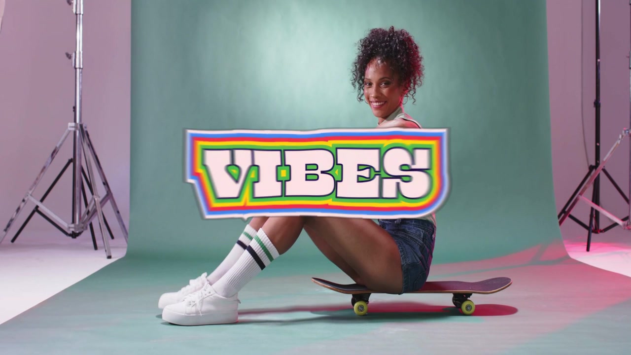 Donna in posa su uno skateboard in uno studio verde menta, che indossa pantaloncini di jeans con sovrapposizione VIBES.