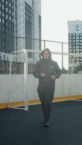 young man jogging in an urban outdoor sports arena with a soccer ball and goalpost in the background, surrounded by high-rise office buildings