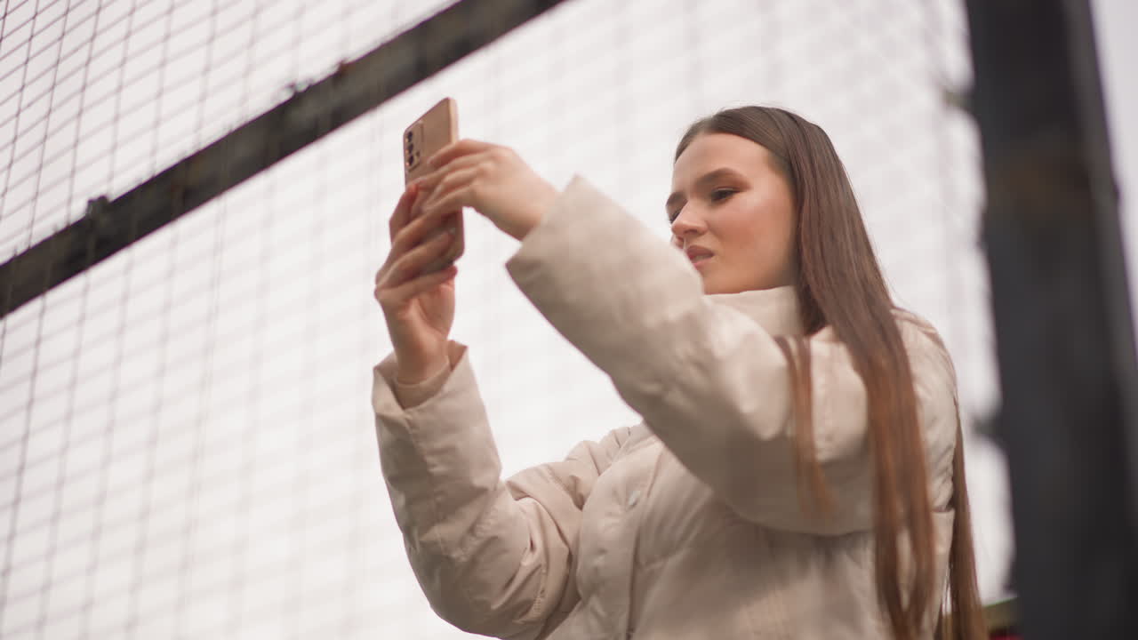 young woman taking selfie outdoors, beige puffer coat and long braided hair, holding smartphone against chainlink fence, testing angles and expressions, posing for social feed, creating portrait