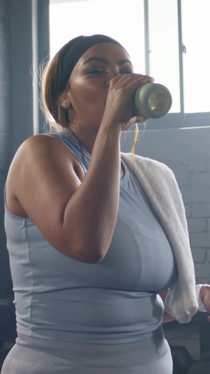 Video verticale: Donna con canotta blu che si idrata a metà allenamento da uno shaker in palestra, asciugandosi con un asciugamano.