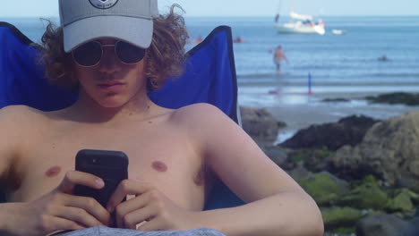 curly-haired man sitting and using smartphone at coverack beach in cornwall, uk