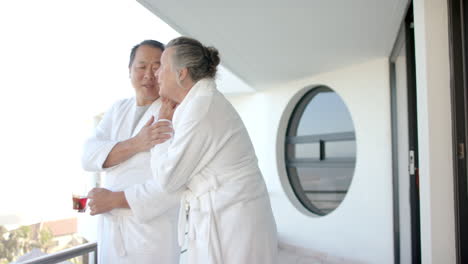 senior couple in bathrobes enjoying balcony view and holding drinks