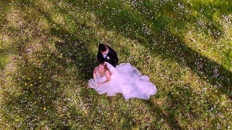 A bride in white gown and groom in tuxedo walk through a peaceful grassy meadow sprinkled with flowers and dappled in long shadows from surrounding spring trees