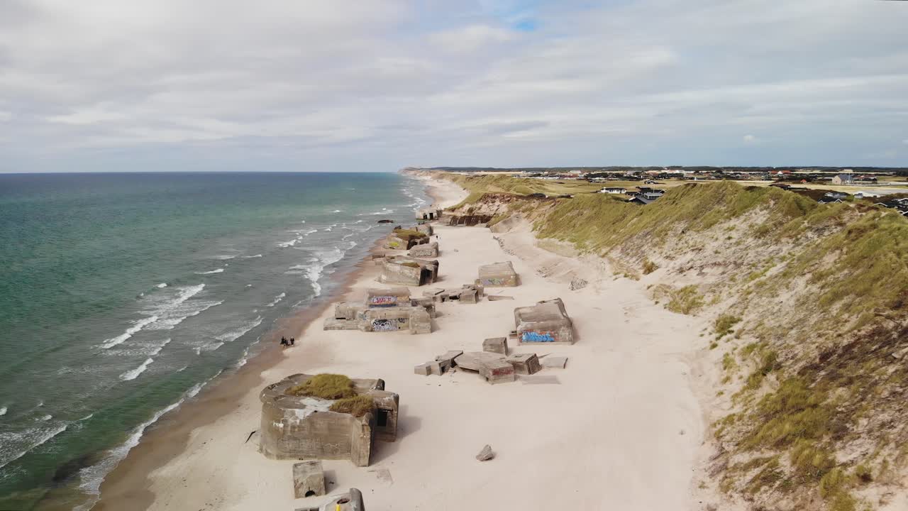 Carrellata aerea in allontanamento di una lunga spiaggia di sabbia bianca sulla costa danese, con diversi vecchi bunker tedeschi abbandonati nella sabbia in una giornata estiva nuvolosa