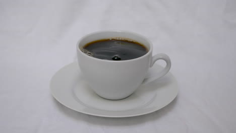 Pouring-sugar-into-white-coffee-cup-with-coffee-on-white-saucer-with-white-background