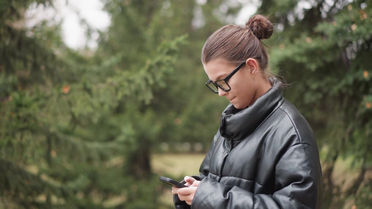 Närbild på en kvinna med glasögon som håller i och tittar på en smartphone i en utomhusparkmiljö. Kvinnan bär en svart läderjacka, hennes hår är uppsatt i en knut, vilket skapar mjuk belysning i scenen.