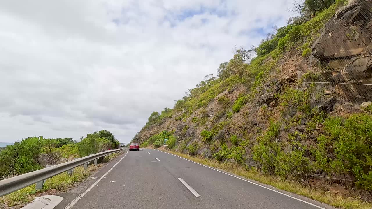 A car navigates the winding Great Ocean Road, surrounded by lush greenery and rocky cliffs under a cloudy sky