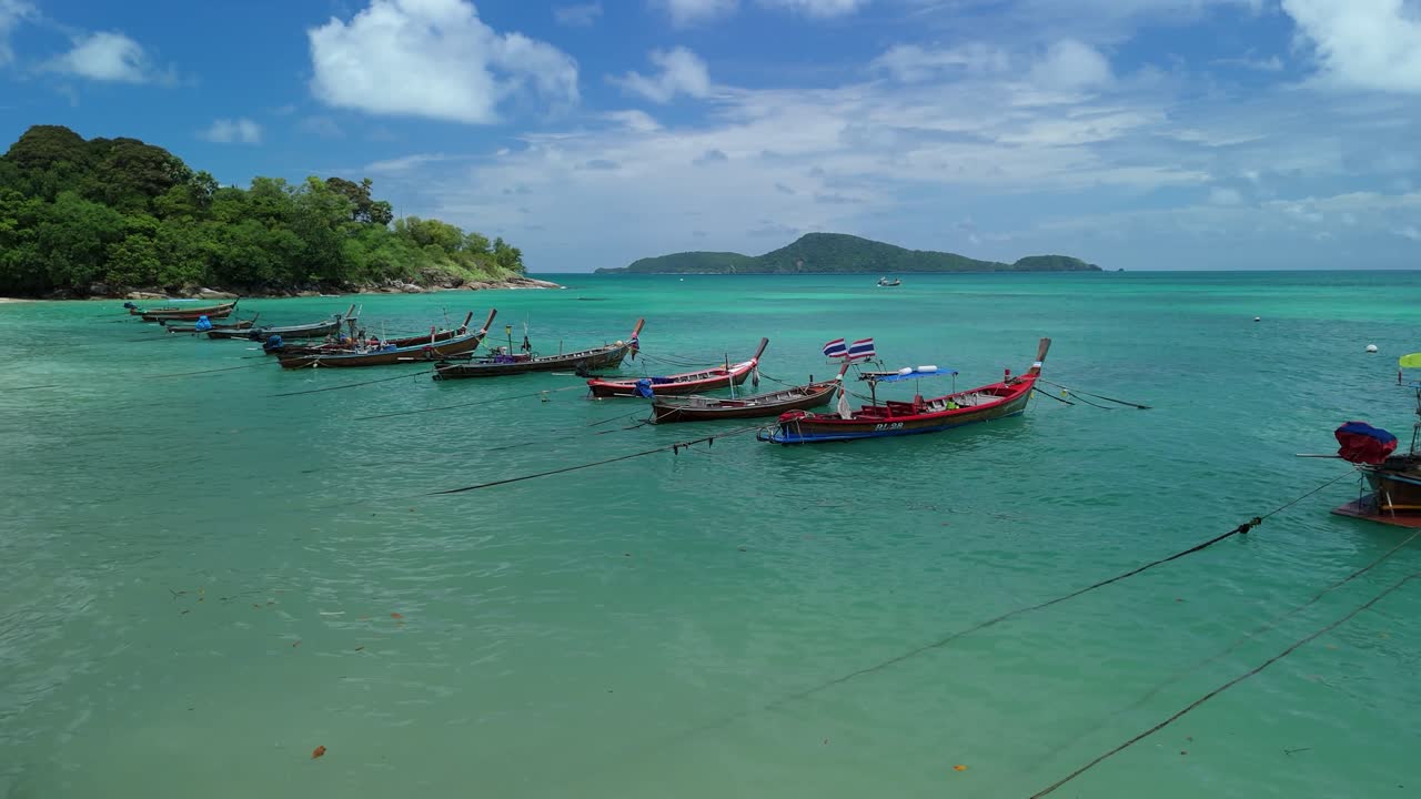 Drone ascends, flying over longtail boats moored along turquoise Rawai Beach
