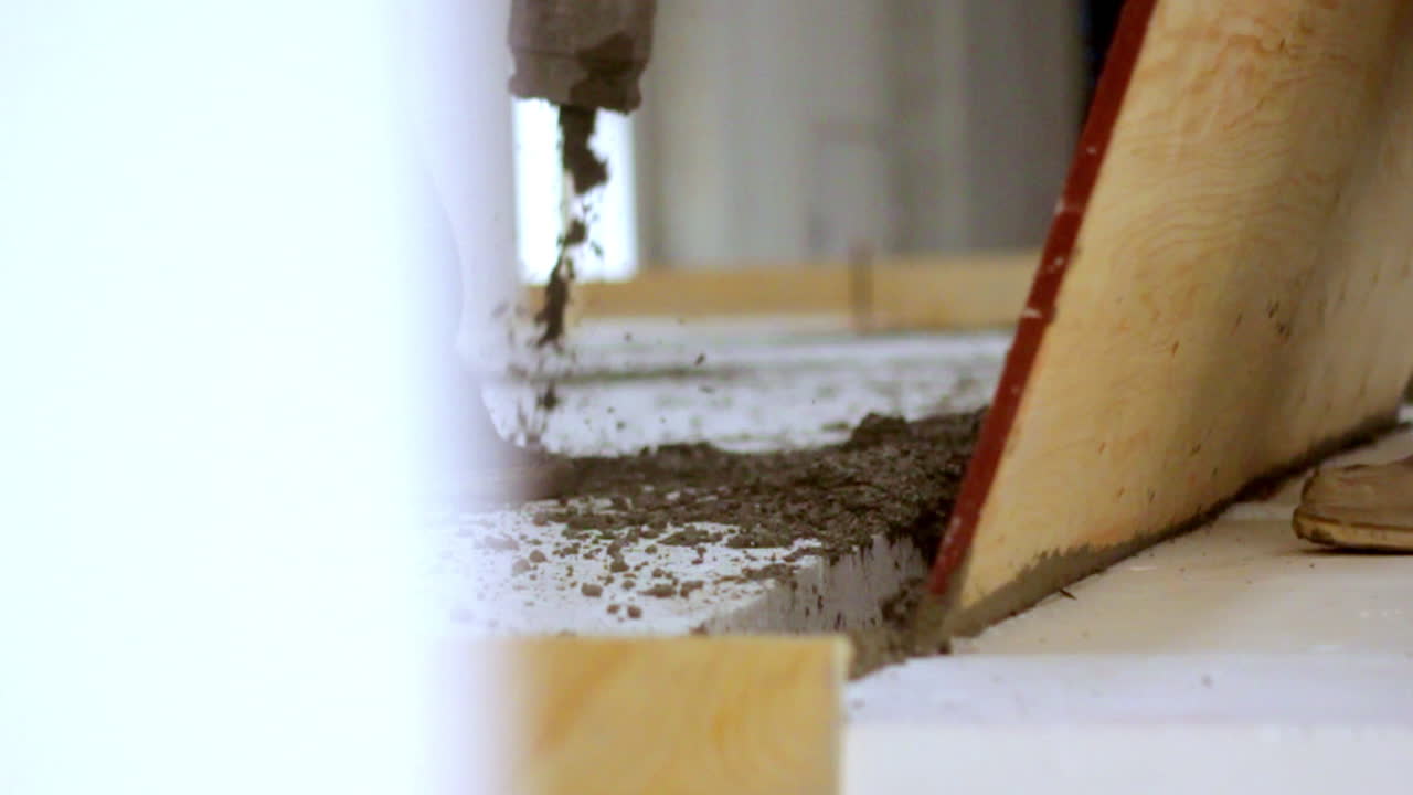 Close-up of a worker leveling fresh concrete inside an industrial building using a wooden screed board. Visible debris, boots, and bright indoor lighting enhance the construction atmosphere