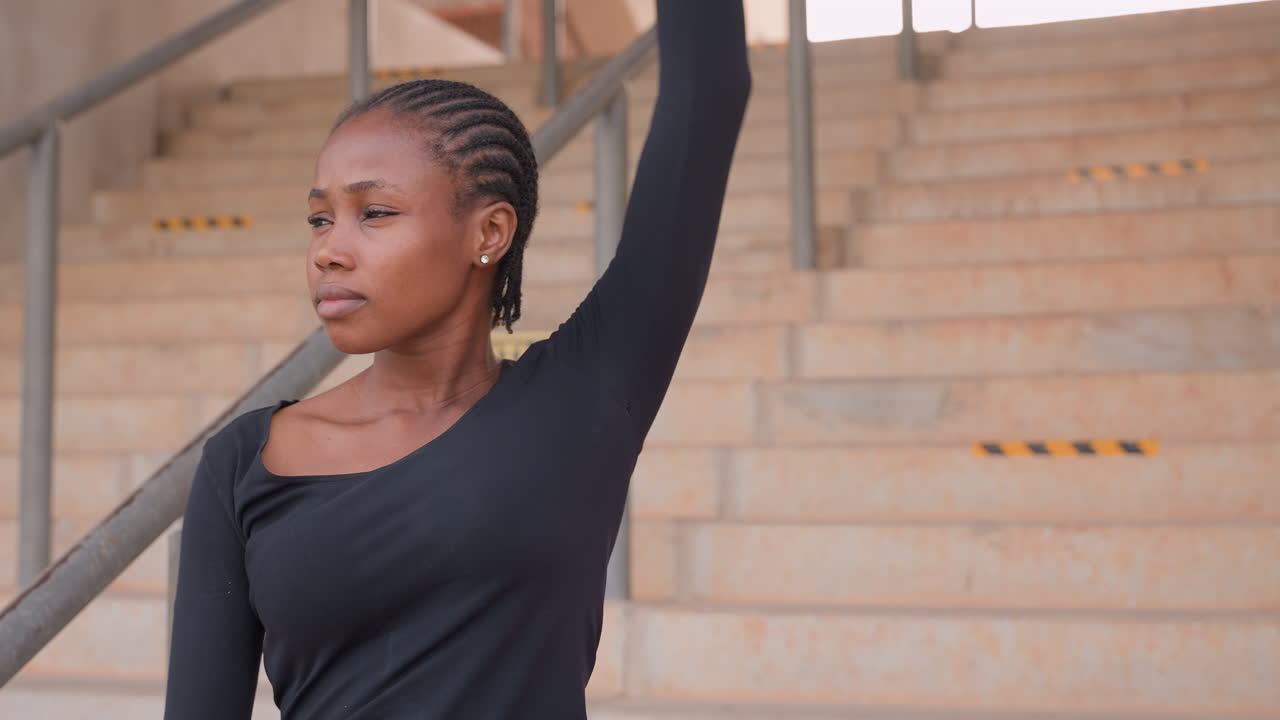 woman on stadium steps demonstrating arm stretch fitness instructor leads movement sequence, steady gaze, raised arm and rhythmic repetition, textured concrete and metal railing create focused urban