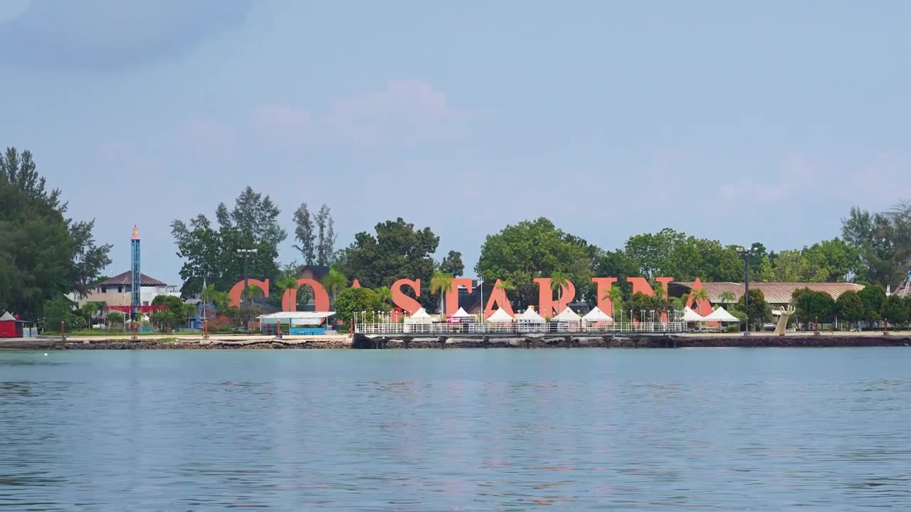 Panoraamakuva suuresta oranssista Coastarina-kyltistä Indonesian Batamin rannikolla, taustalla trooppisia puita ja valkoisia telttoja. Ihanteellinen matkailu- ja aluuutisille