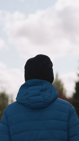 vista posterior cercana de una persona con chaqueta azul y gorra negra paseando por un área residencial, con casas y árboles borrosos en el fondo bajo un cielo nublado