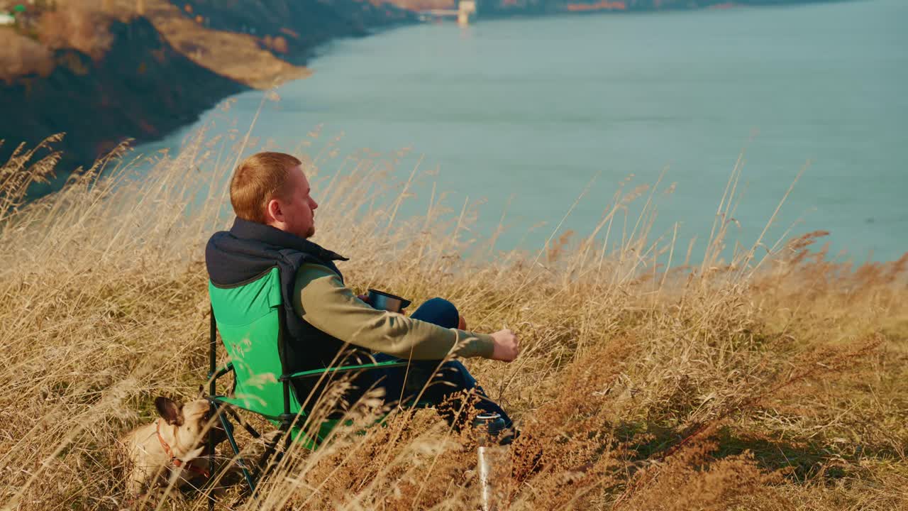 Un momento di tranquillità nella natura: uomo seduto su una sedia su una collina che guarda un lago sereno mentre si gode una bevanda calda con il suo cane al suo fianco, circondato da erba alta e viste panoramiche