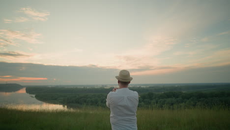 a thoughtful man wearing a white shirt, hat, and jeans stands in a grassy field by a tranquil lake at sunset, gazing into the distance. the warm sunset light create a peaceful atmosphere