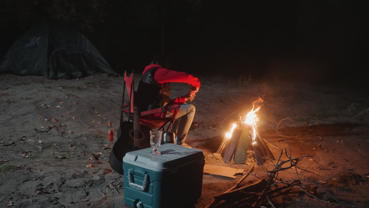 Natcamping med en kaukasisk camper, der passer bålet. En rød klapstol og køleboks står ved siden af en guitar på jorden, mens brænde og optændingsmateriale arrangeres. En varm glød oplyser den grusbelagte plads, med teltets silhuet i baggrunden, i stilhed.